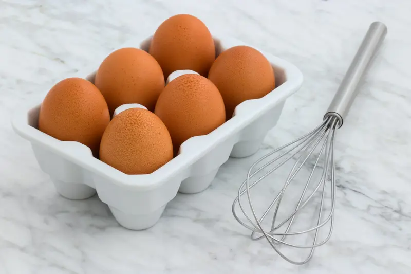 A carton of eggs beside a whisk